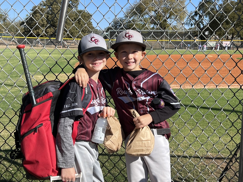 Two young Rough Riders baseball teammates smiling together beside the field
